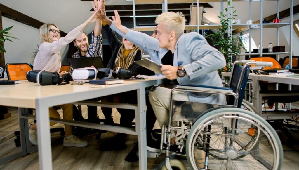 Happy multiethnic diverse young people sitting in office and giving high five each other after great collaboration, discussing strategies. Disabled male manager sitting with his colleagues at office